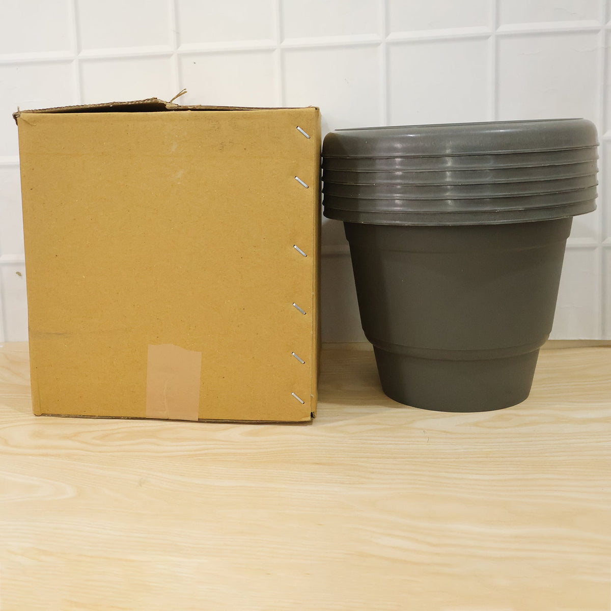 A brown cardboard box next to a stack of five gray plastic flower pots on a light wooden surface.