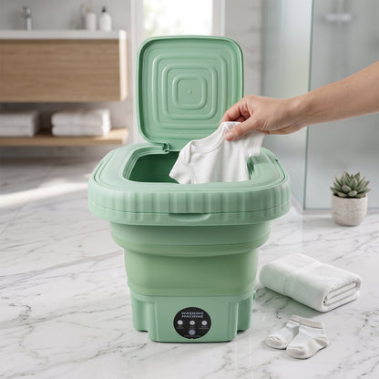 A green portable washing machine with its lid open, showing water swirling inside, with folded white clothes beside it in a