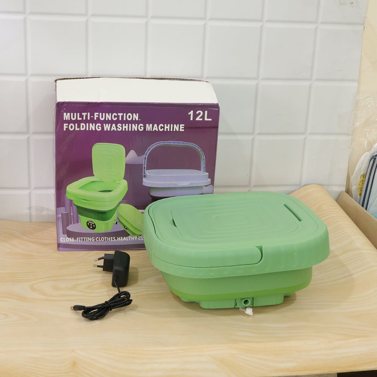 A green portable washing machine, collapsed with its lid closed, sits next to its product box and power adapter on a wooden