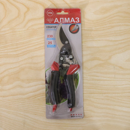 Red and black garden pruners in clear plastic and red cardboard packaging on a light wood background.
