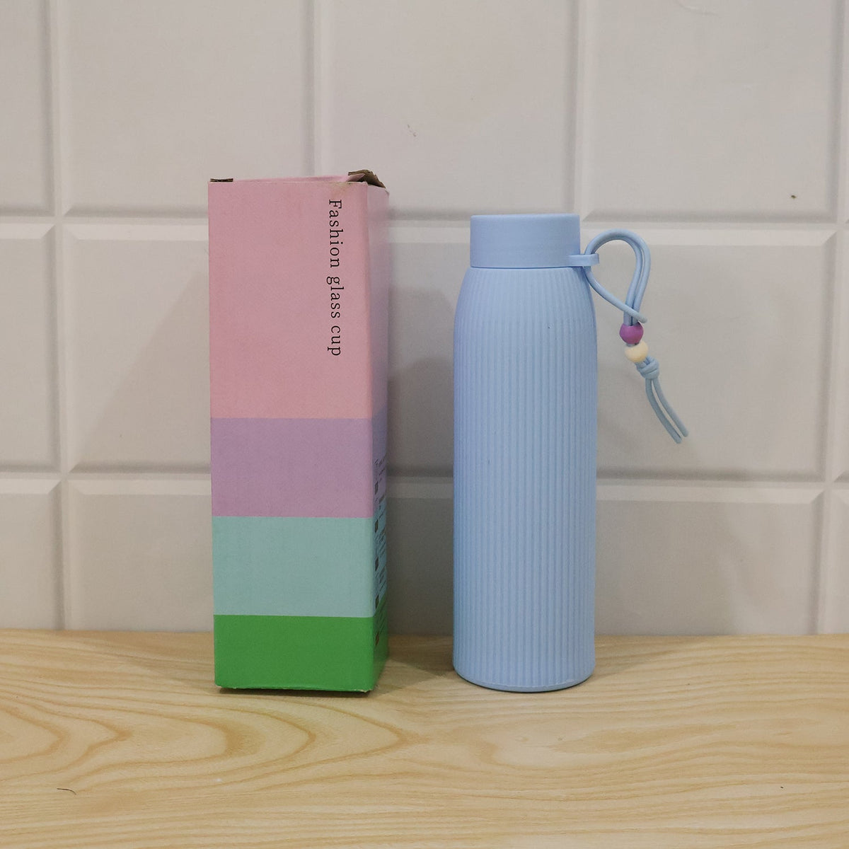 A light blue ribbed glass water bottle with a beaded strap sits next to a colorful pink, blue, and green box labeled