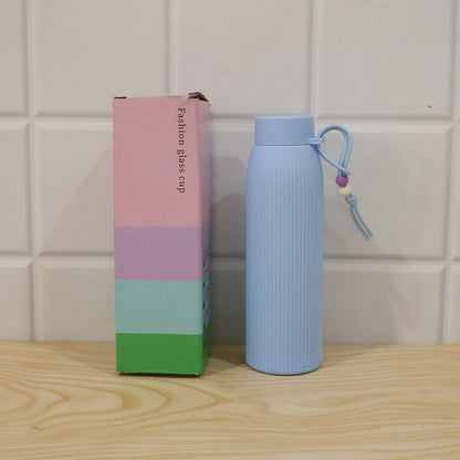 A light blue ribbed glass water bottle with a beaded strap sits next to a colorful pink, blue, and green box labeled