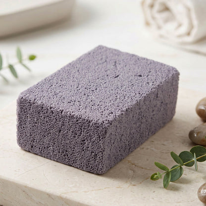 A single gray cleaning block with a porous texture resting on a light marble surface with green leaves and pebbles.