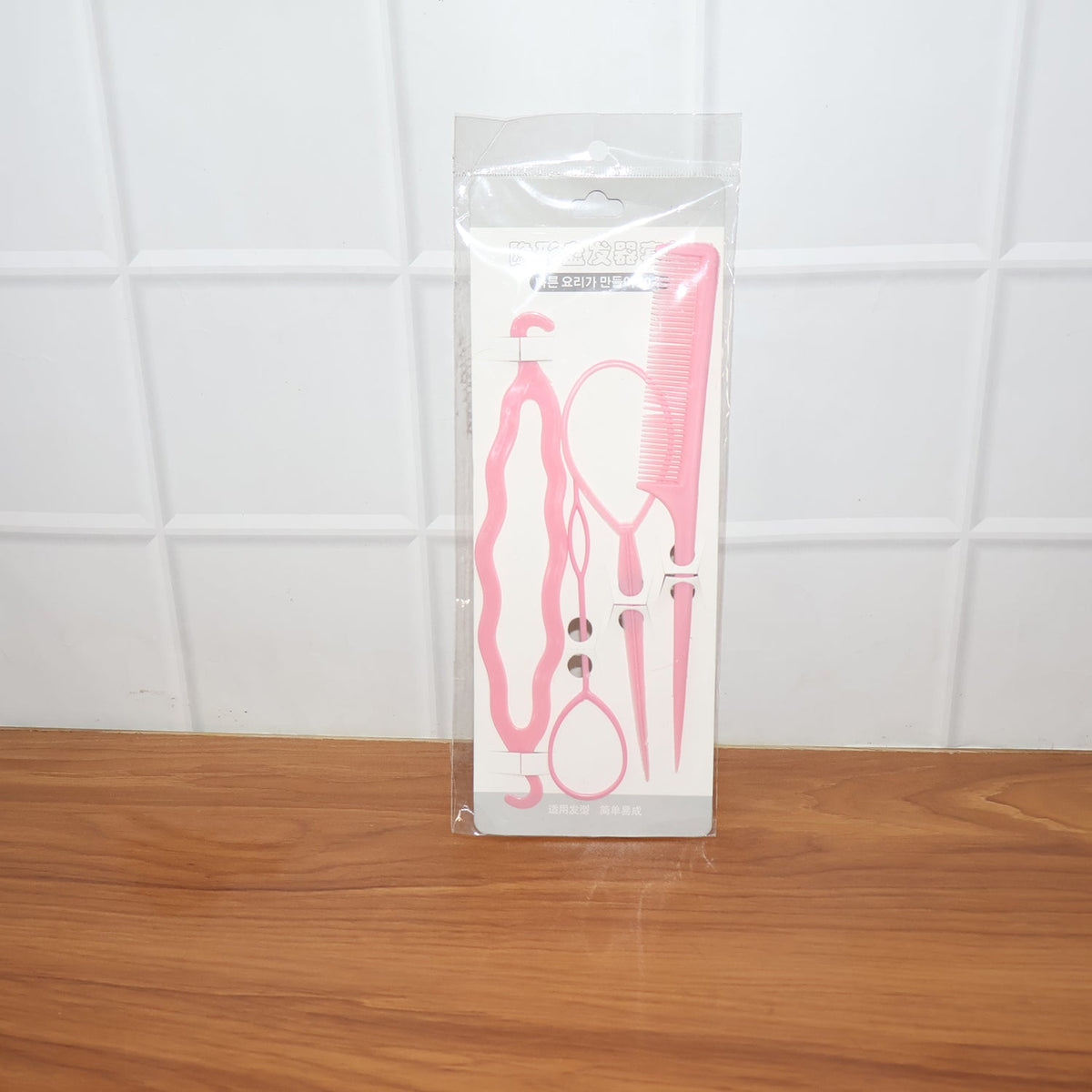 A clear plastic package containing four pink hair styling tools stands upright on a wooden surface against a white tiled