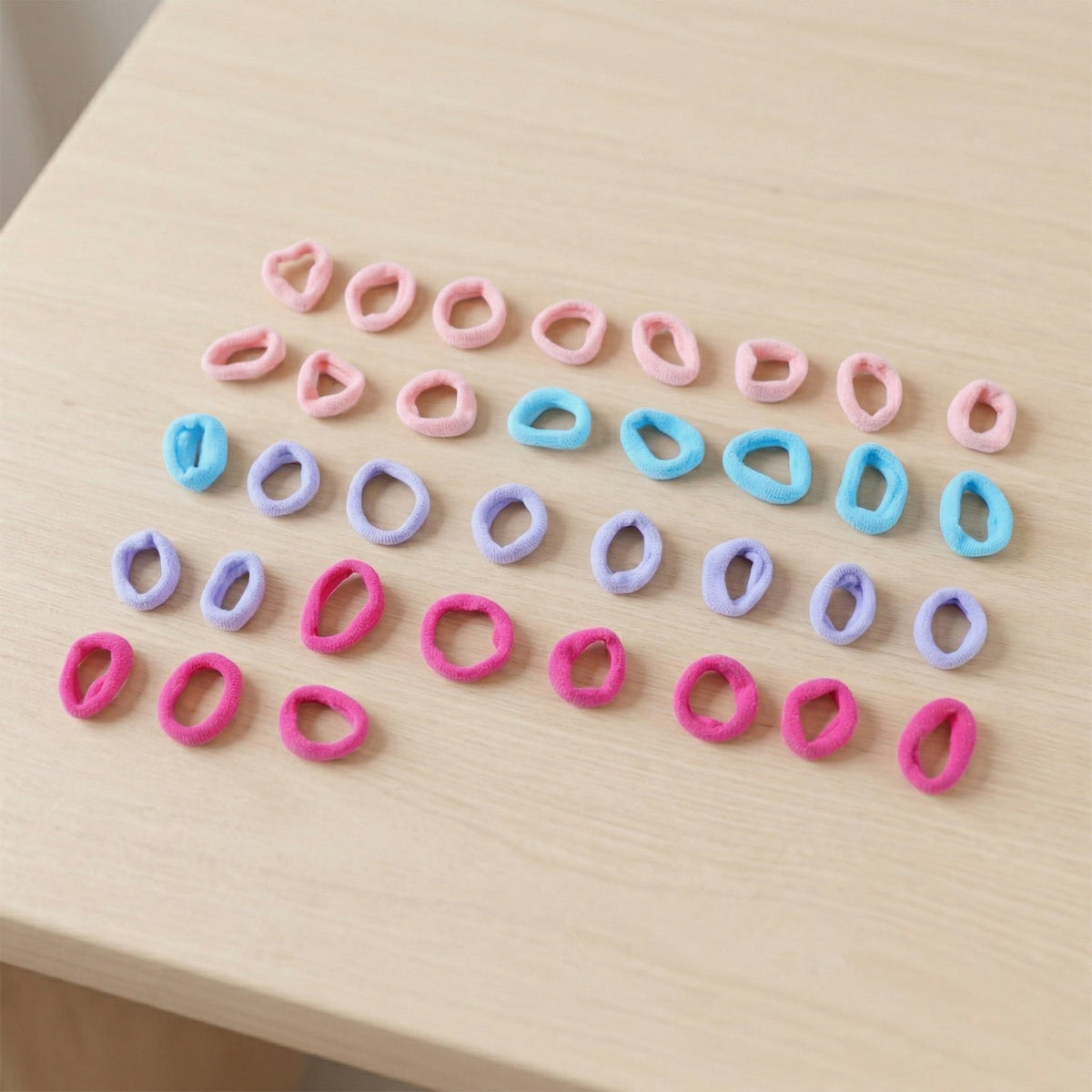 Thirty-five assorted pink, blue, lavender, and fuchsia elastic hair ties scattered on a light wooden surface.