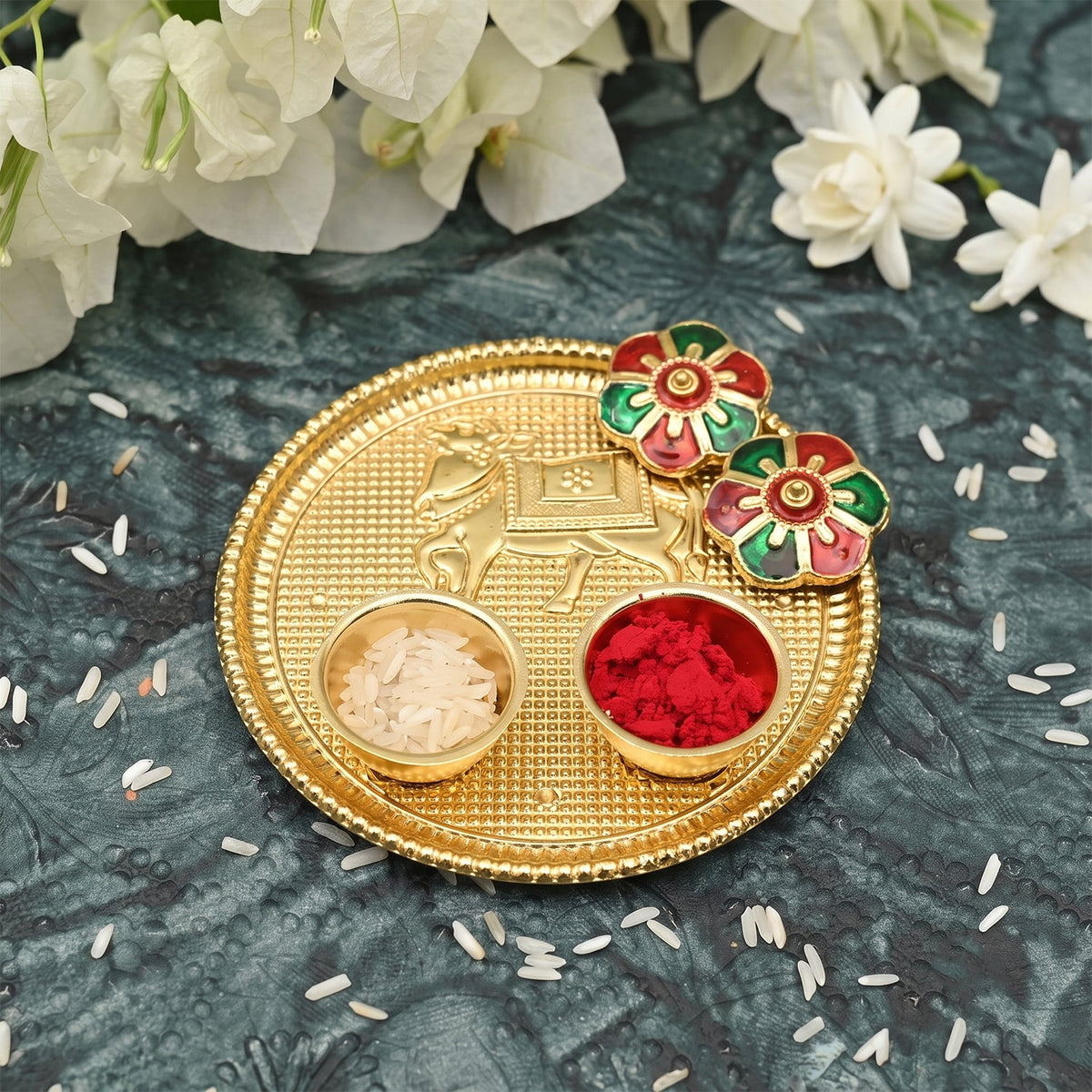 A golden pooja thali with a cow design, two small bowls of rice and kumkum, and two flower-shaped lids on a dark patterned