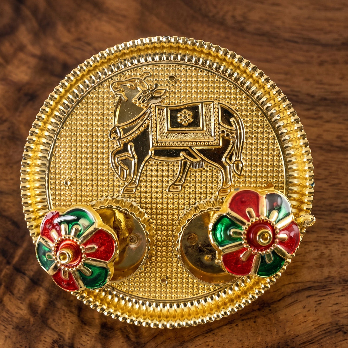 A close-up of a golden pooja thali with an embossed cow design and two colorful flower-shaped lids on a wooden surface.