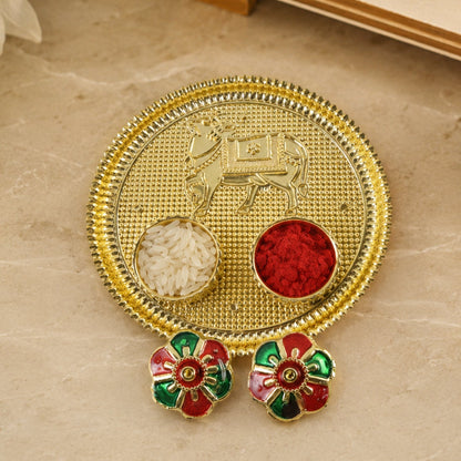 A golden pooja thali with a cow design, two small bowls of rice and kumkum, and two colorful flower-shaped lids on a light