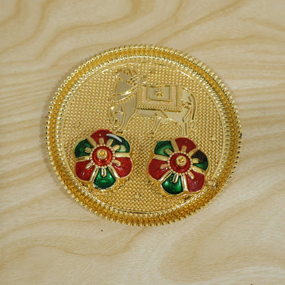 A golden pooja thali with an embossed cow design and two colorful flower-shaped lids on a light wooden background.