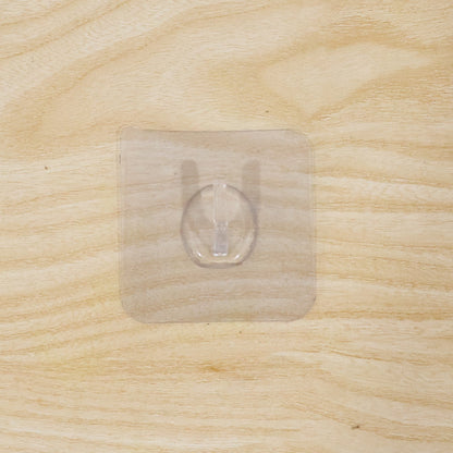 A clear self-adhesive wall hook centered on a light, unfinished wooden surface with a visible grain.