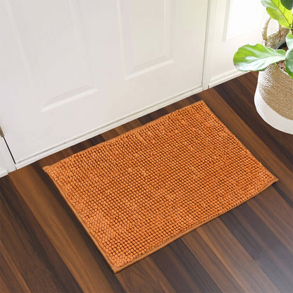 An orange textured bobble mat rests on a dark wooden floor in front of a white door, beside a potted plant.