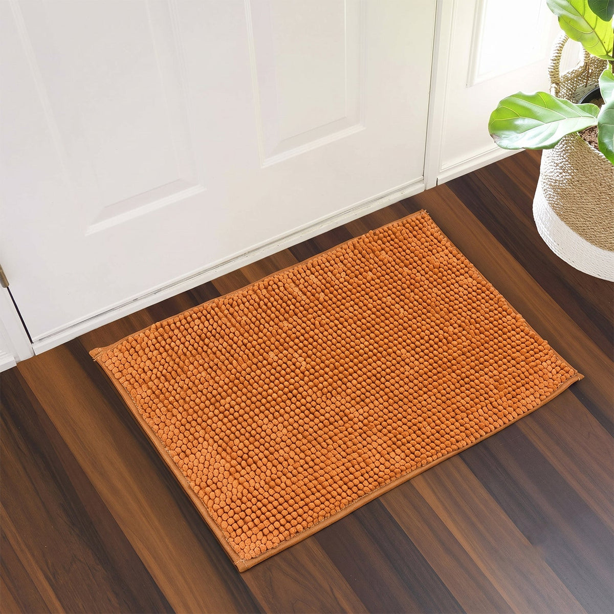 An orange textured bobble mat rests on a dark wooden floor in front of a white door, beside a potted plant.