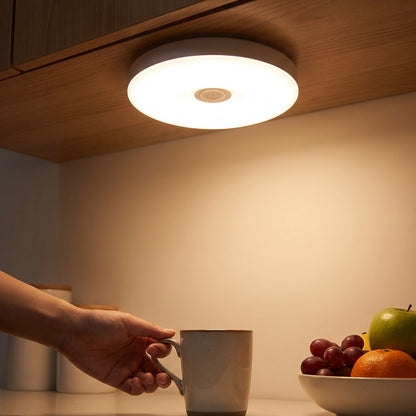 A hand reaches for a mug under a bright, round LED light mounted beneath a wooden cabinet, illuminating a fruit bowl