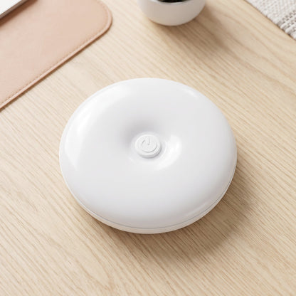A white, round LED light with a central power button rests on a light wood desk next to a pink mat