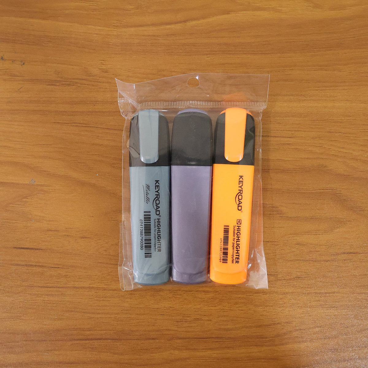 Three Keyroad Metallic highlighters in light blue, purple, and orange are sealed in clear plastic packaging on a wooden