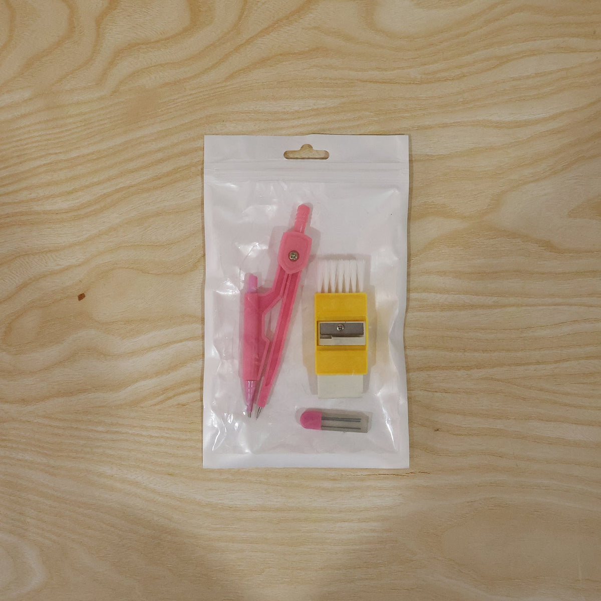 The pink compass, yellow eraser with brush, and lead refills are sealed in clear plastic packaging on a light wooden surface.
