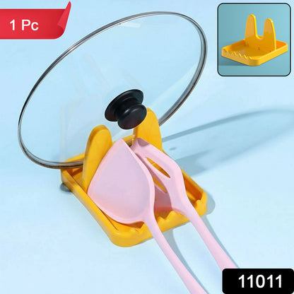 A yellow plastic lid and spoon rest holding a glass pot lid and two pink silicone spatulas, with a small inset image of the