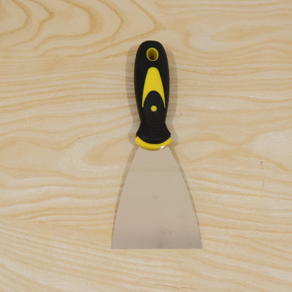 A stainless steel putty knife with a black and yellow handle lies flat on a light-colored wooden surface.