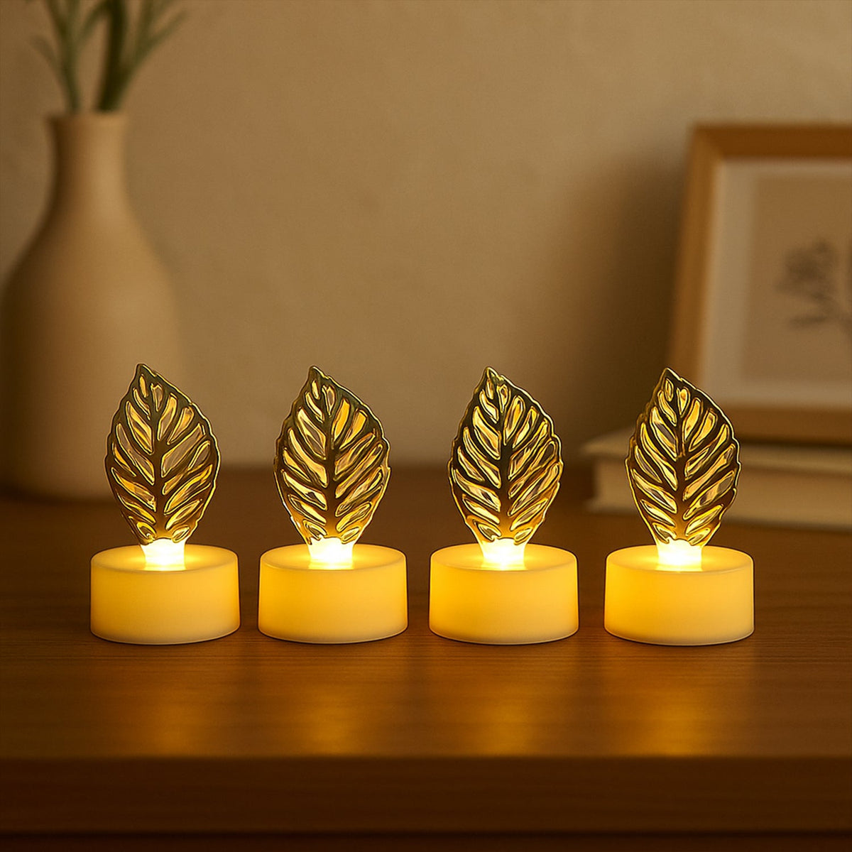 Four golden leaf LED candles sit on a wooden surface with a light-colored vase and framed art in the background.