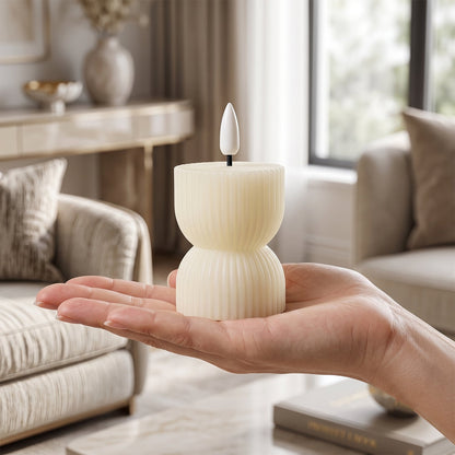 A hand holds one cream ribbed flameless candle with a white wick, centered in a softly lit living room setting.