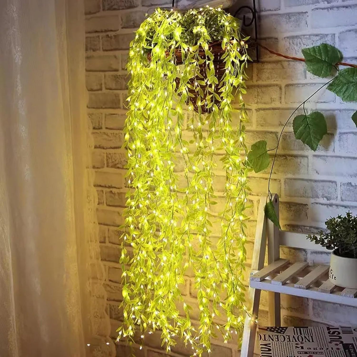 A hanging basket with long strands of green leaf string lights cascading down a brick wall, illuminated, with a small potted