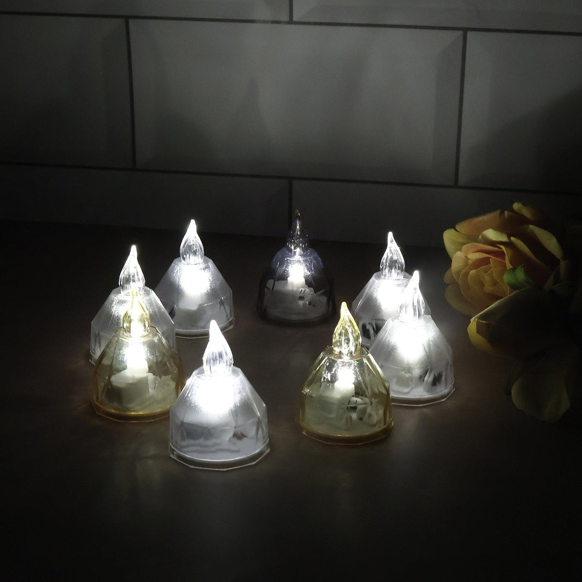 Seven glowing LED diyas, a mix of clear and amber, arranged on a dark surface with a blurred flower in the background.