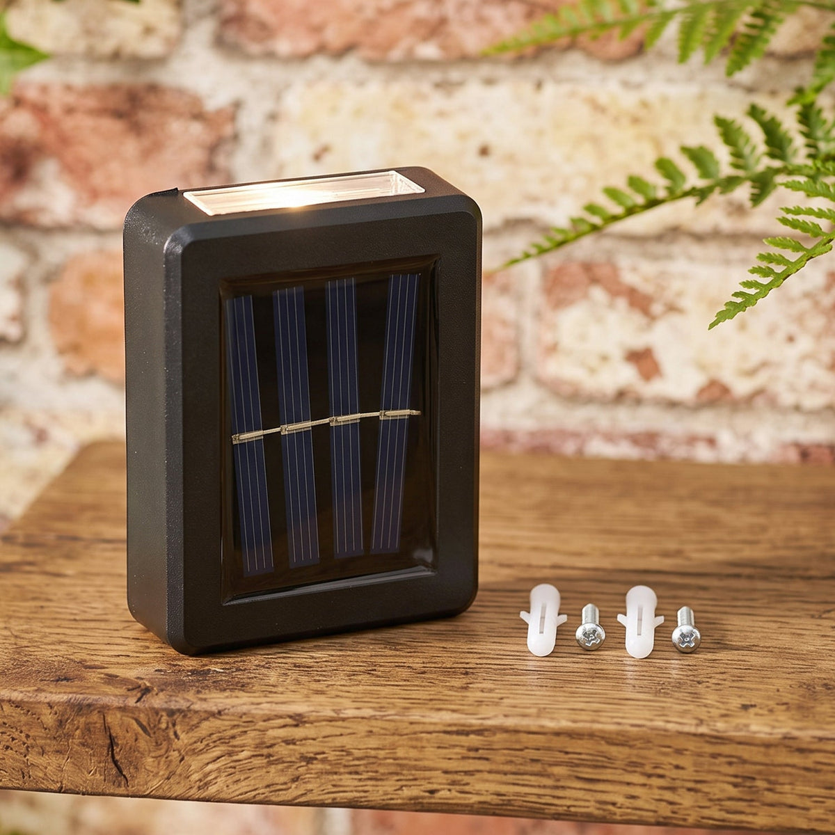 A black solar LED wall light with a blue solar panel sits on a wooden surface next to two white wall plugs and two silver