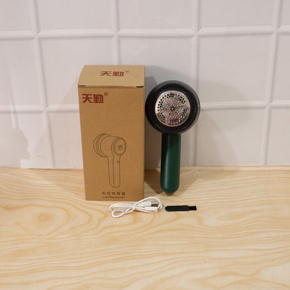 A dark green and black lint remover, its brown box, a white USB-C cable, and a small black brush are arranged on a light