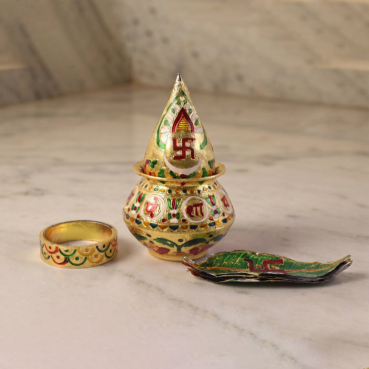 A golden metal Kalash with a pointed top, a ring, and a leaf-shaped dish on marble.