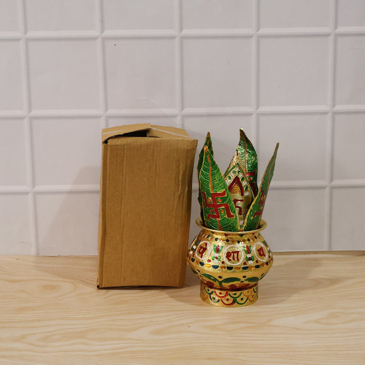 A golden metal Kalash with green leaves next to a brown cardboard box on a wooden surface.