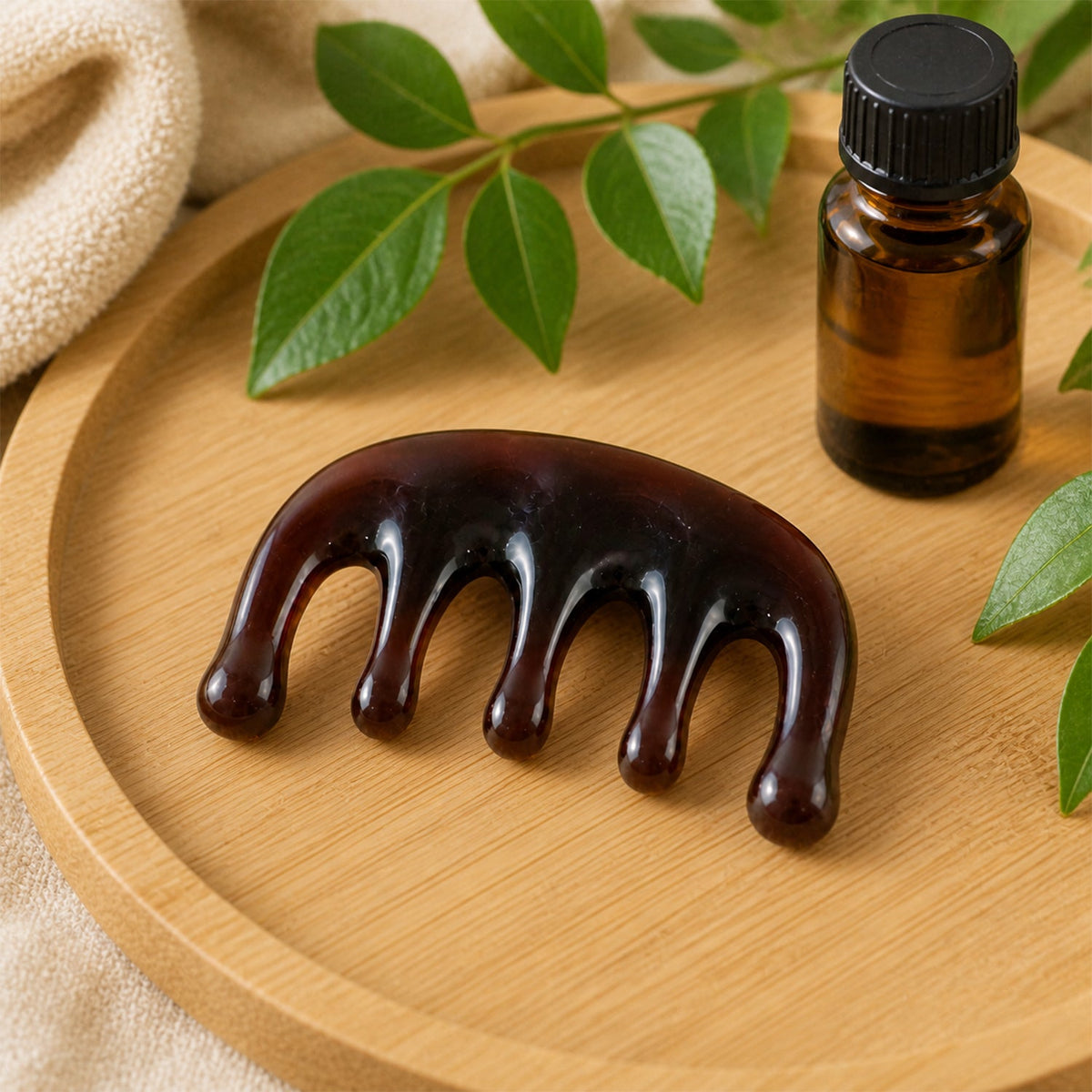A dark brown resin scalp massager rests on a light wooden tray next to a small amber essential oil bottle and green leaves.