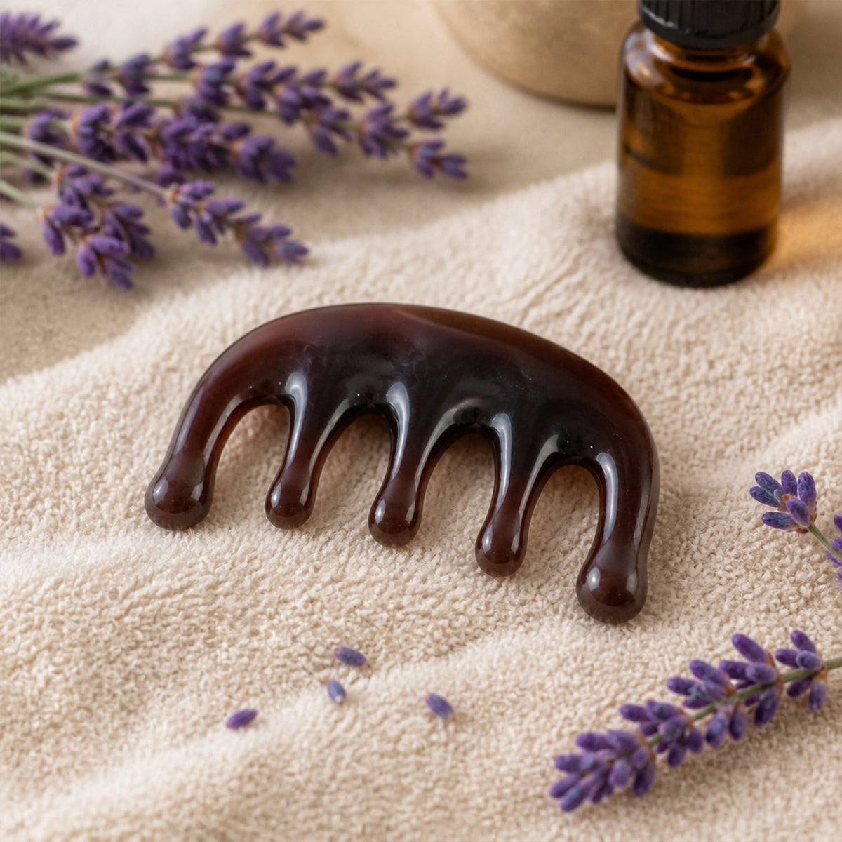 A dark brown resin scalp massager lies on a textured beige towel, surrounded by sprigs of purple lavender and an amber