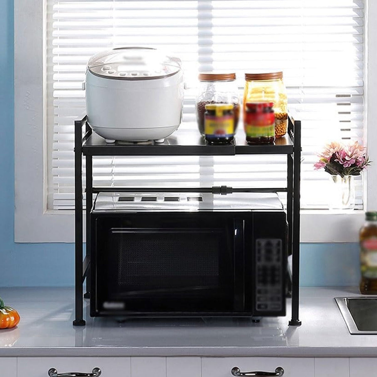 Black microwave on a black metal rack, with a rice cooker and jars on top, by a window with blinds.