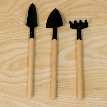 Two small black trowels and a matching rake with light wooden handles are neatly arranged on a wooden surface.