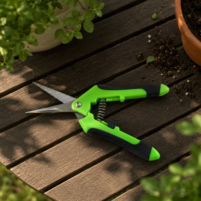 Green and black snips rest open on a wooden table next to potted plants and scattered soil.