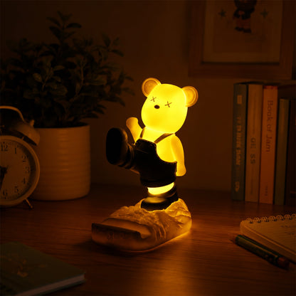 A glowing yellow bear phone holder with dark overalls on a desk at night, next to books.