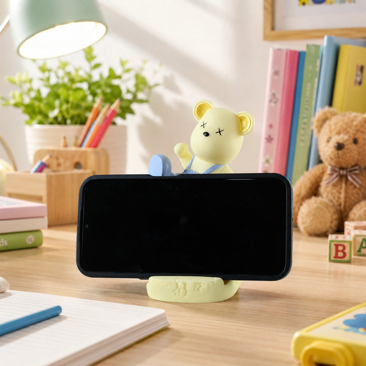A yellow bear phone holder with blue overalls on a desk in daylight, holding a phone horizontally.