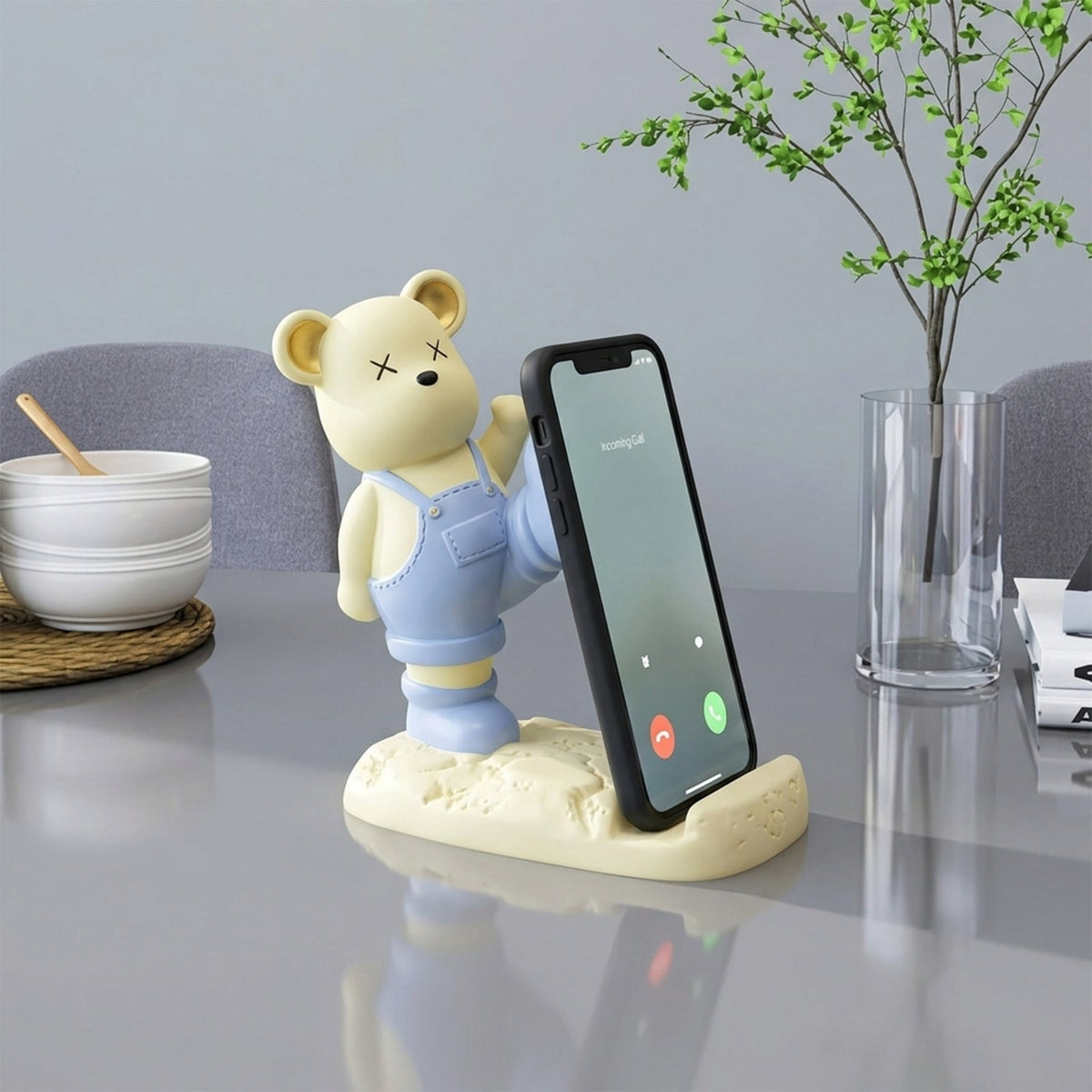 A yellow bear phone holder with blue overalls on a grey table, holding a phone vertically.