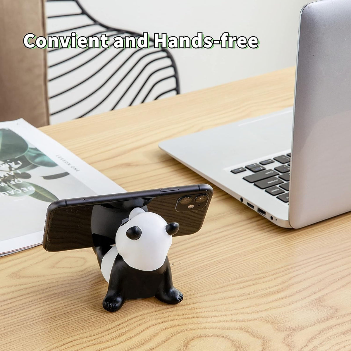 A black and white panda figurine holds a black smartphone horizontally on a wooden desk next to a laptop.