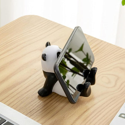 A black and white panda figurine holds a black smartphone on a wooden desk, viewed from behind the phone.