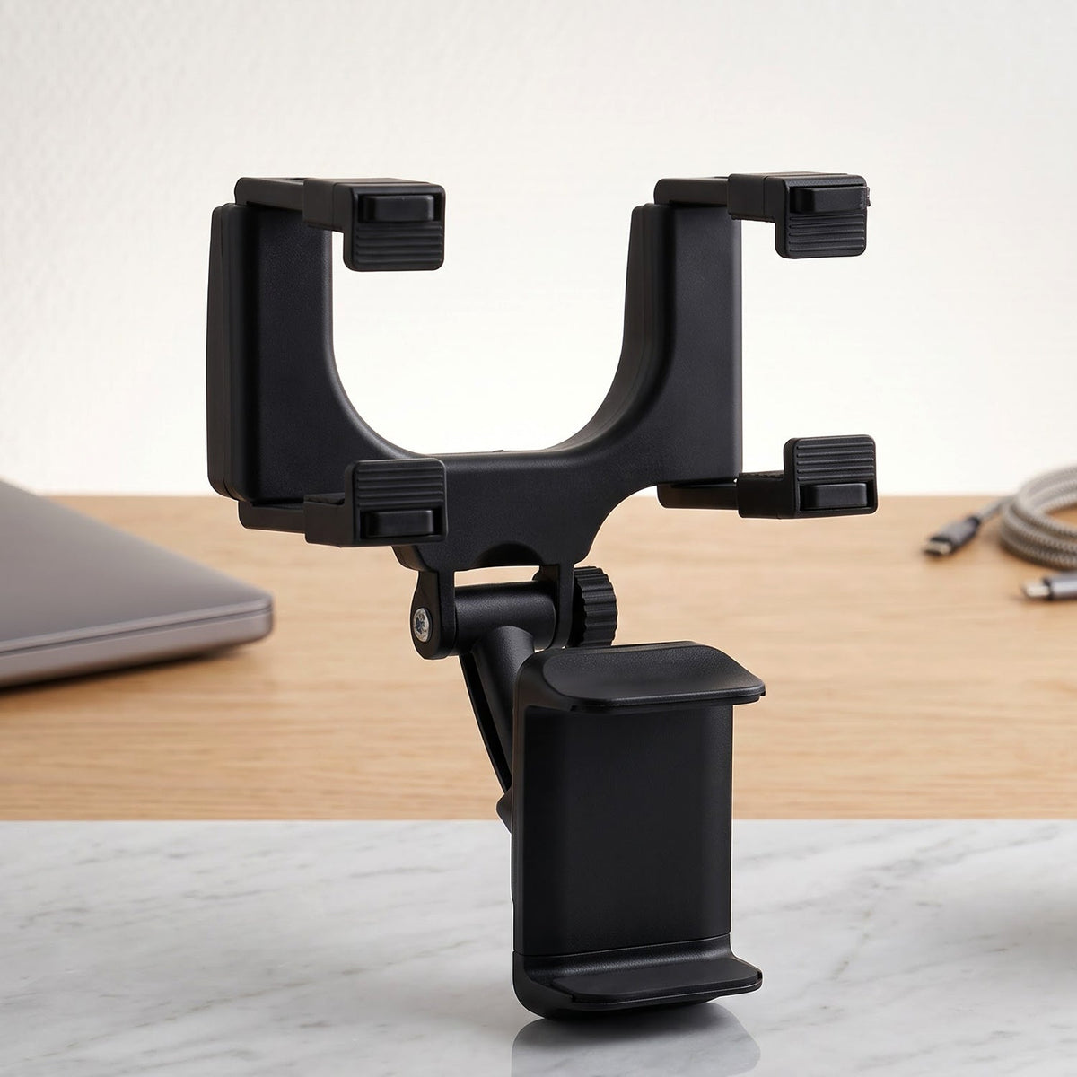 A black car phone mount on a wooden desk, with a laptop and charging cable in the background.