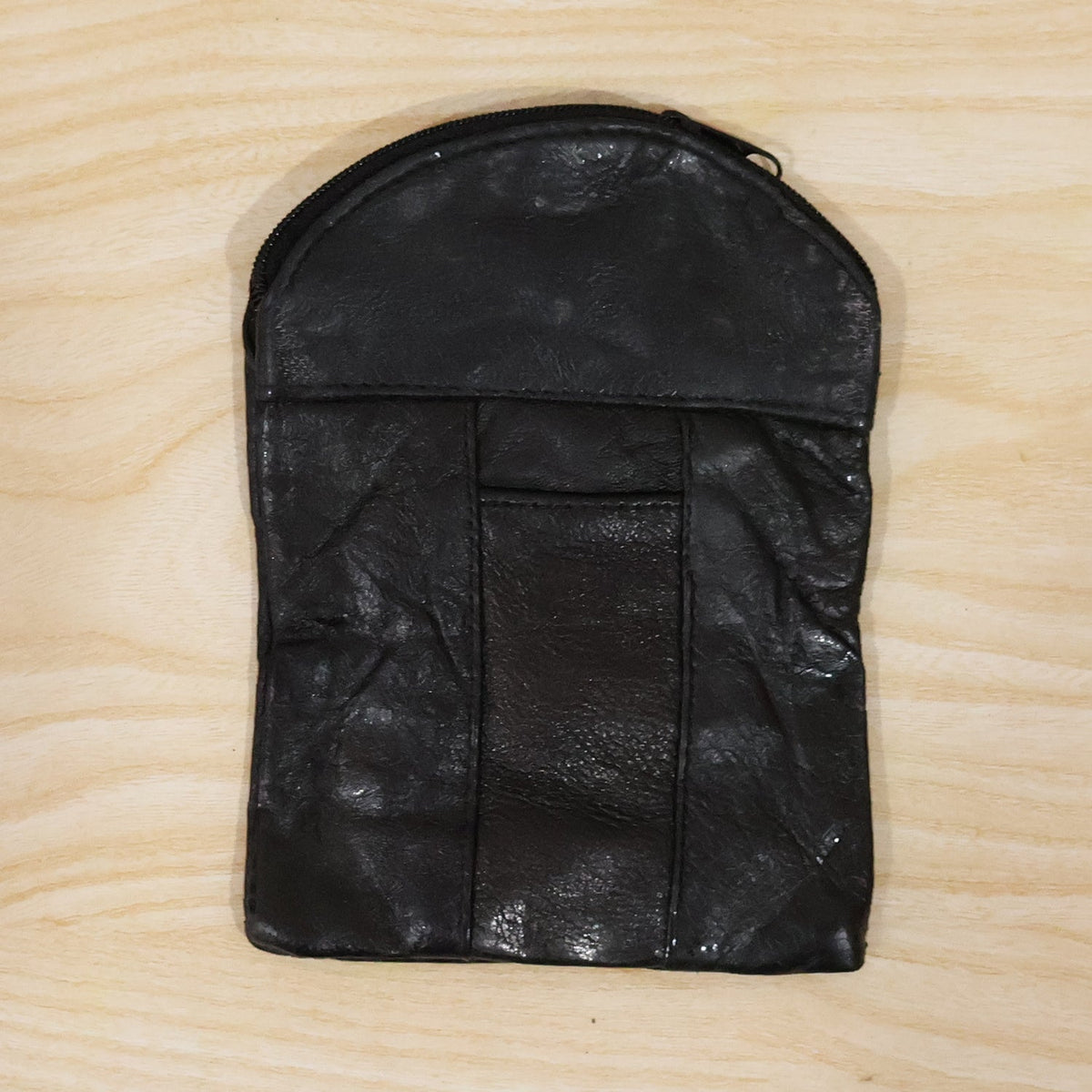 A black leather pouch with a top zipper and vertical seam details, laid flat on a light wooden surface.