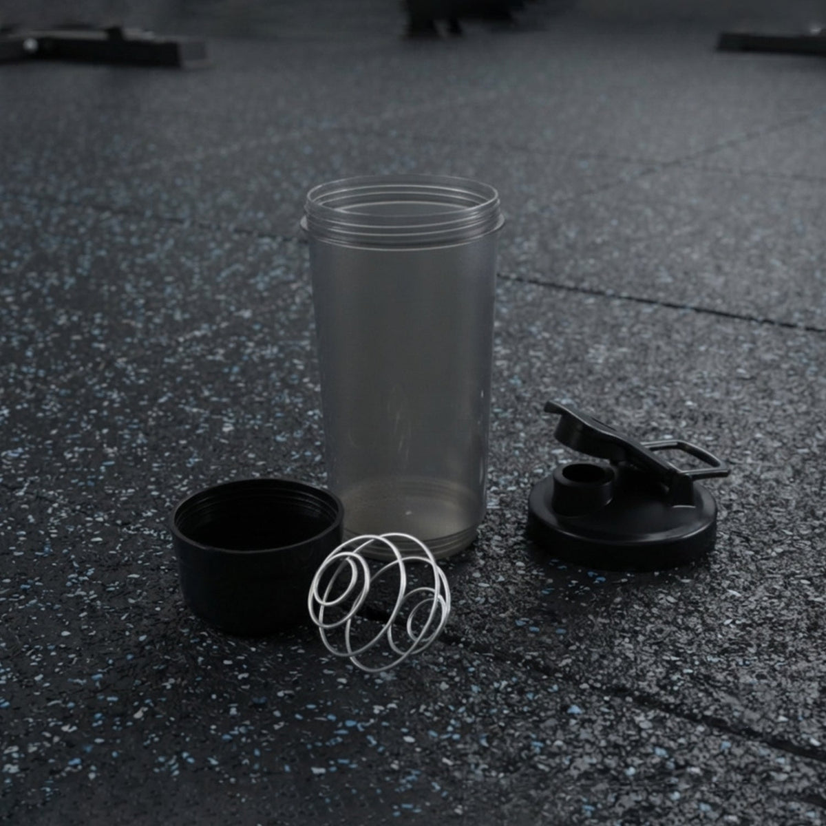 A disassembled grey and black shaker bottle with a metal whisk is on a dark gym floor.