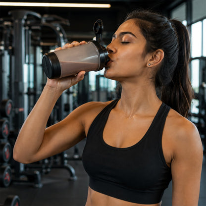 A woman in a black sports bra drinks from a grey and black shaker bottle in a gym.