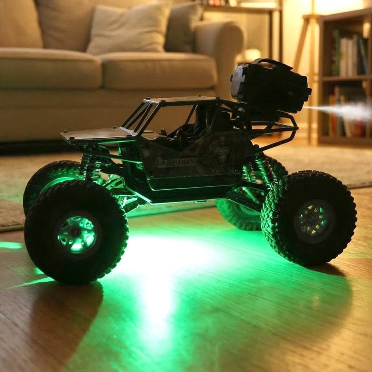 A black remote control rock crawler with green glowing wheels and a bright white headlight sits on a wooden floor indoors.