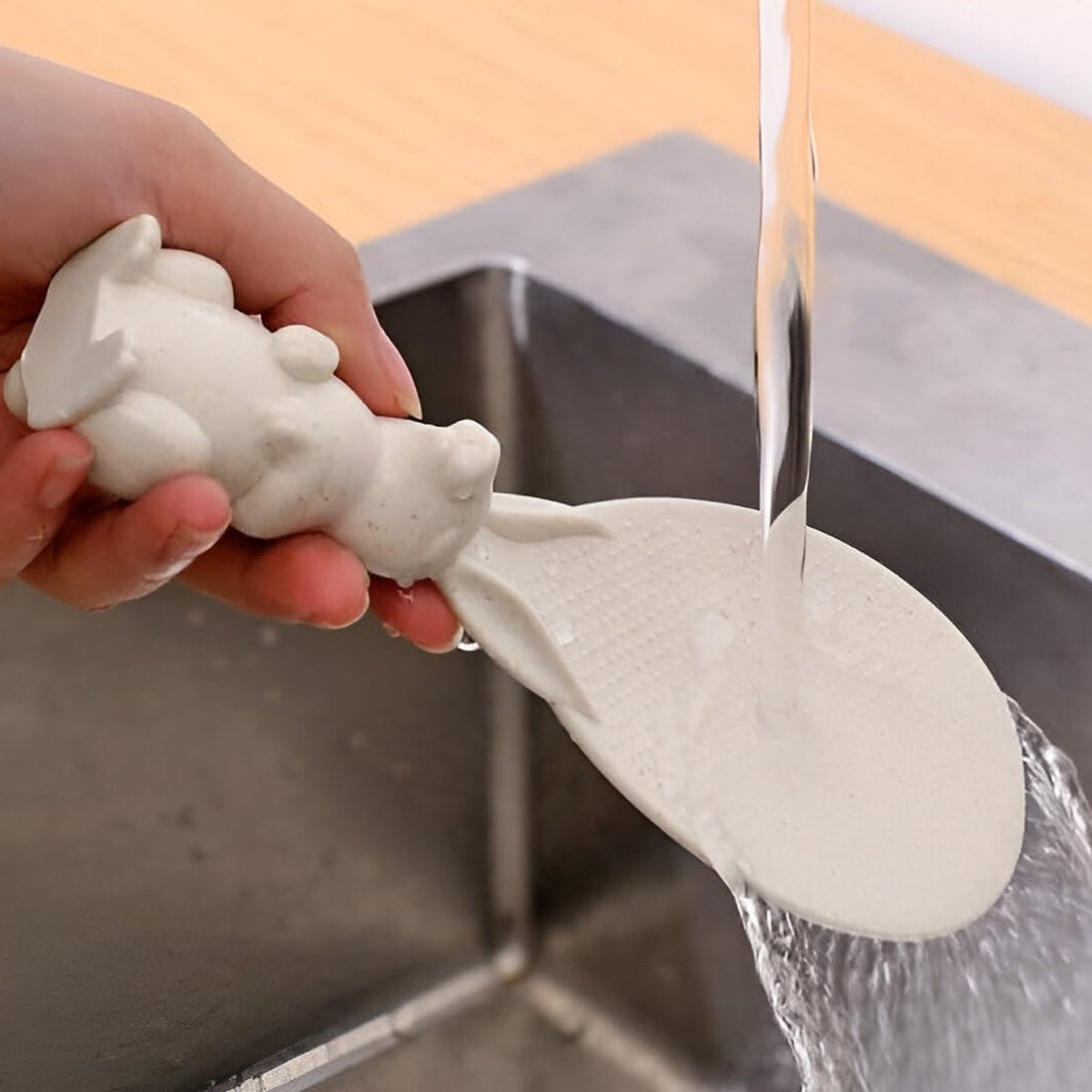 A hand holds a white bunny-handled rice spoon under running water in a stainless steel sink, showing it being washed.