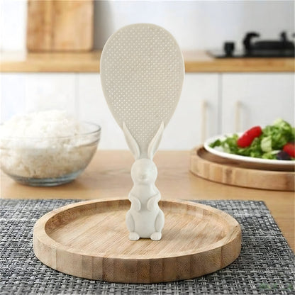 A white bunny-handled rice spoon stands upright on a wooden tray, with a bowl of rice and salad in the background.