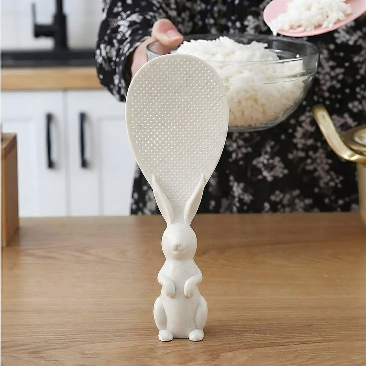 A white bunny-handled rice spoon stands on a wooden counter, with a person holding a bowl of rice in the background.