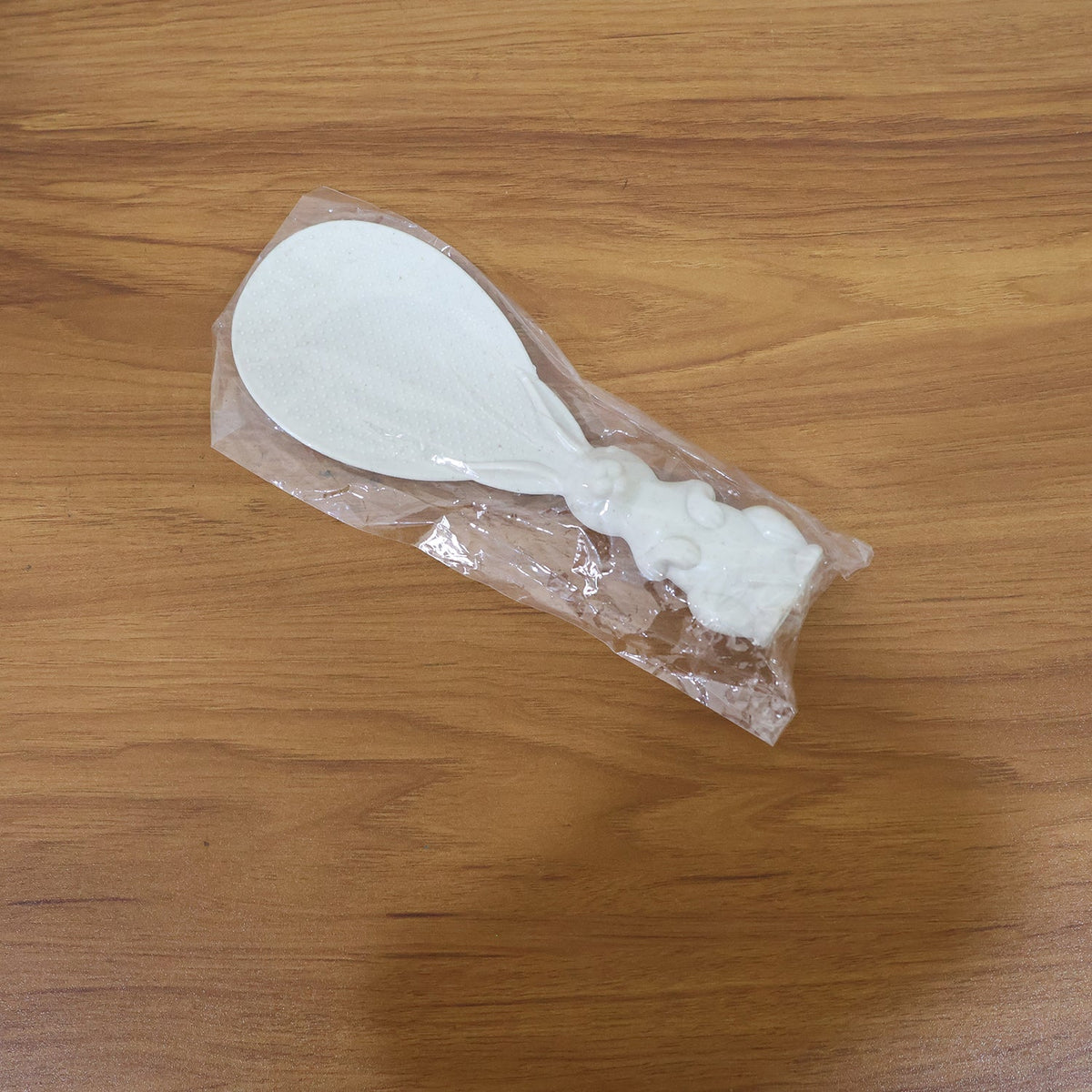 A white bunny-handled rice spoon is sealed in clear plastic packaging, lying on a wooden surface.