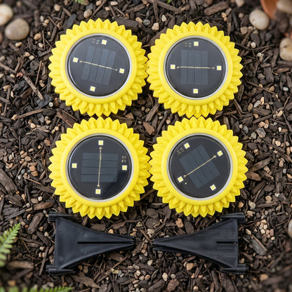 Four yellow, sunflower-shaped solar lights with black solar panels and two black ground stakes are arranged on dark soil.